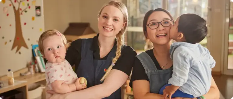 Two educators holding young children in a nursery learning space.