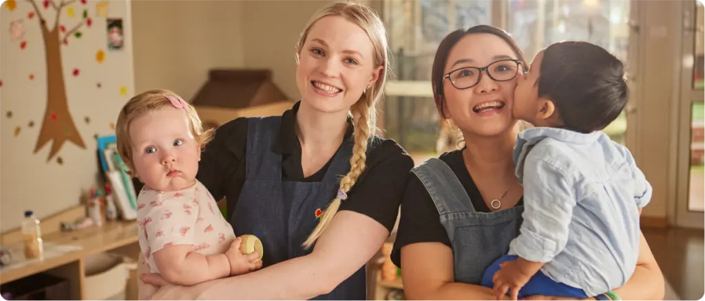 Two educators holding young children in a nursery learning space.